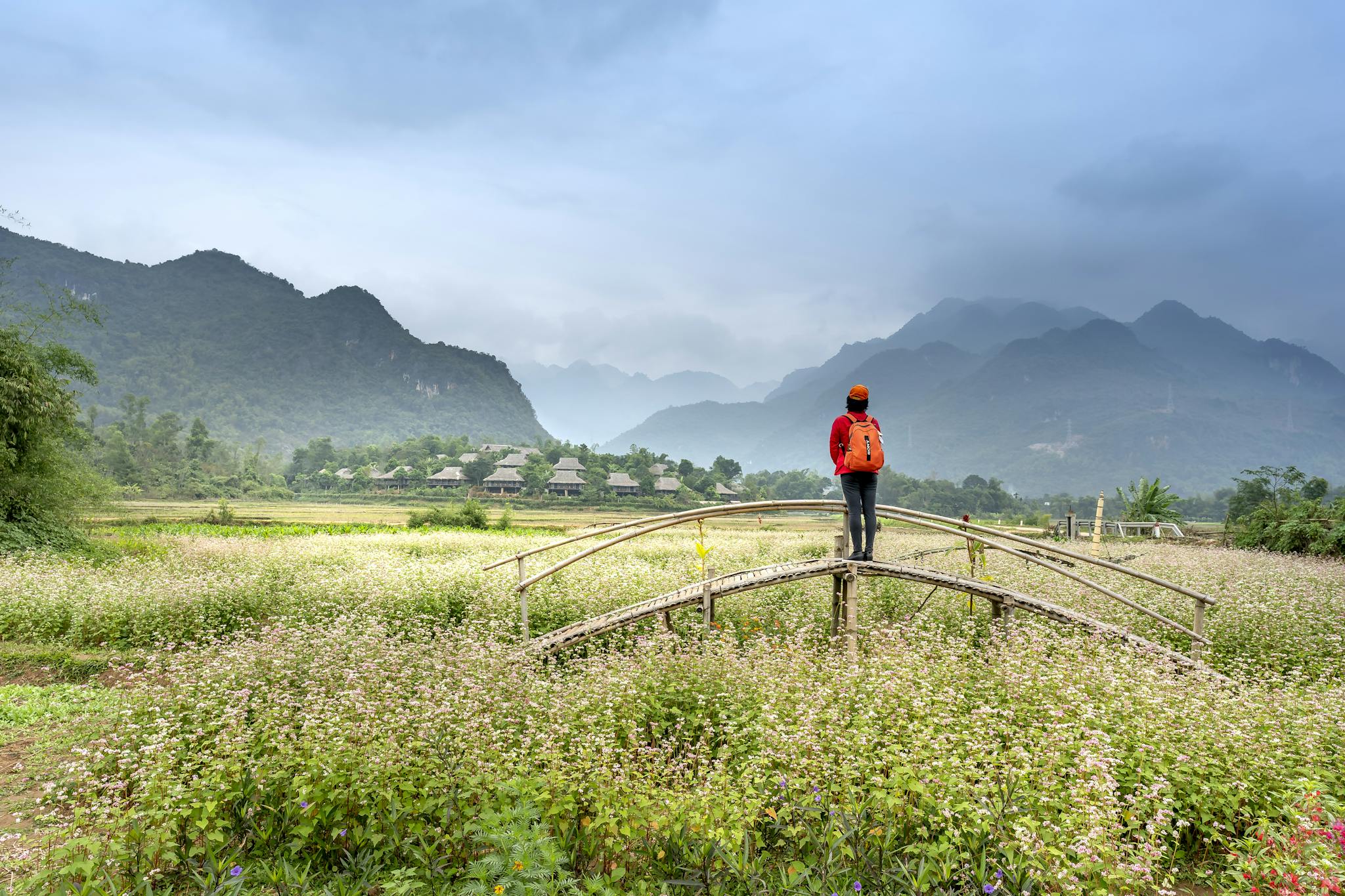 Person with backpack standing on a bridge in a lush valley with mountains and fields.